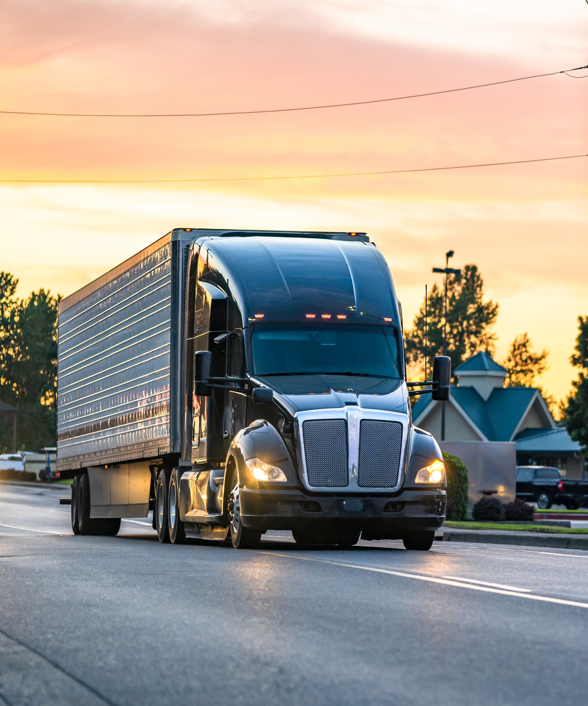 Black high cab big rig semi truck with turned on headlights transporting cargo in refrigerator semi trailer running on the local road at sunset time